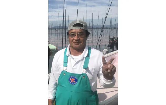 漁師姿の徳永義昭さんが、船の上でピースサインをして微笑んでいる